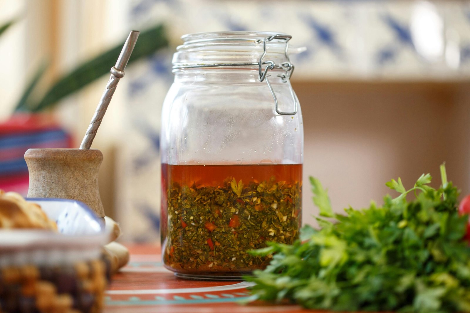 A glass jar with a metal clasp lid filled with chimichurri sauce, placed on a table with fresh parsley, a mate gourd, and a bombilla straw in the background.