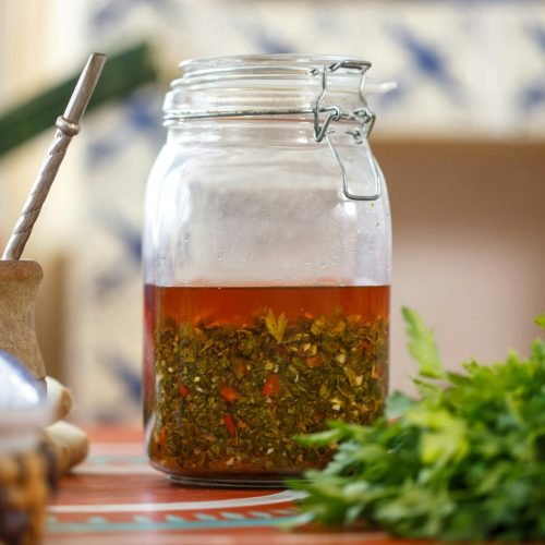 A jar of homemade chimichurri sauce sits on a table, ready to enhance any dish, alongside fresh parsley and a traditional mate setup.